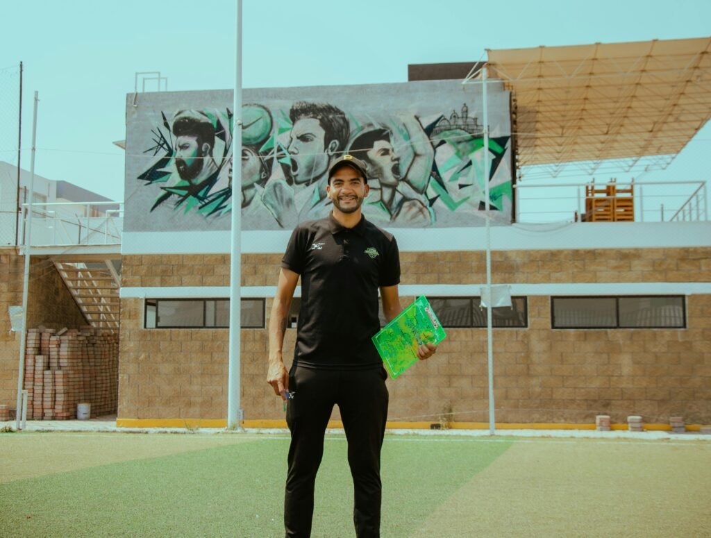 Smiling soccer coach stands on field with sports mural in the background, promoting training and sportsmanship.